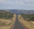 A vehicle driving along a straight sealed highway through dry Australian bush towards rocky hills