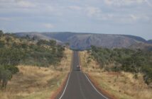 A vehicle driving along a straight sealed highway through dry Australian bush towards rocky hills