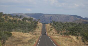 A vehicle driving along a straight sealed highway through dry Australian bush towards rocky hills