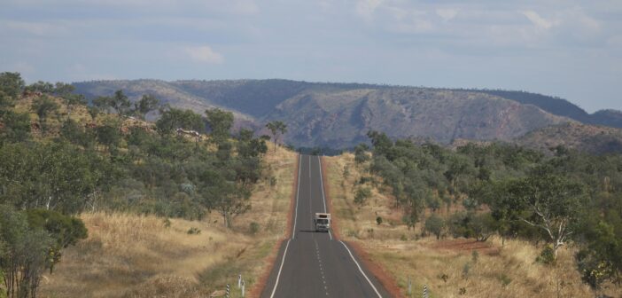 A vehicle driving along a straight sealed highway through dry Australian bush towards rocky hills